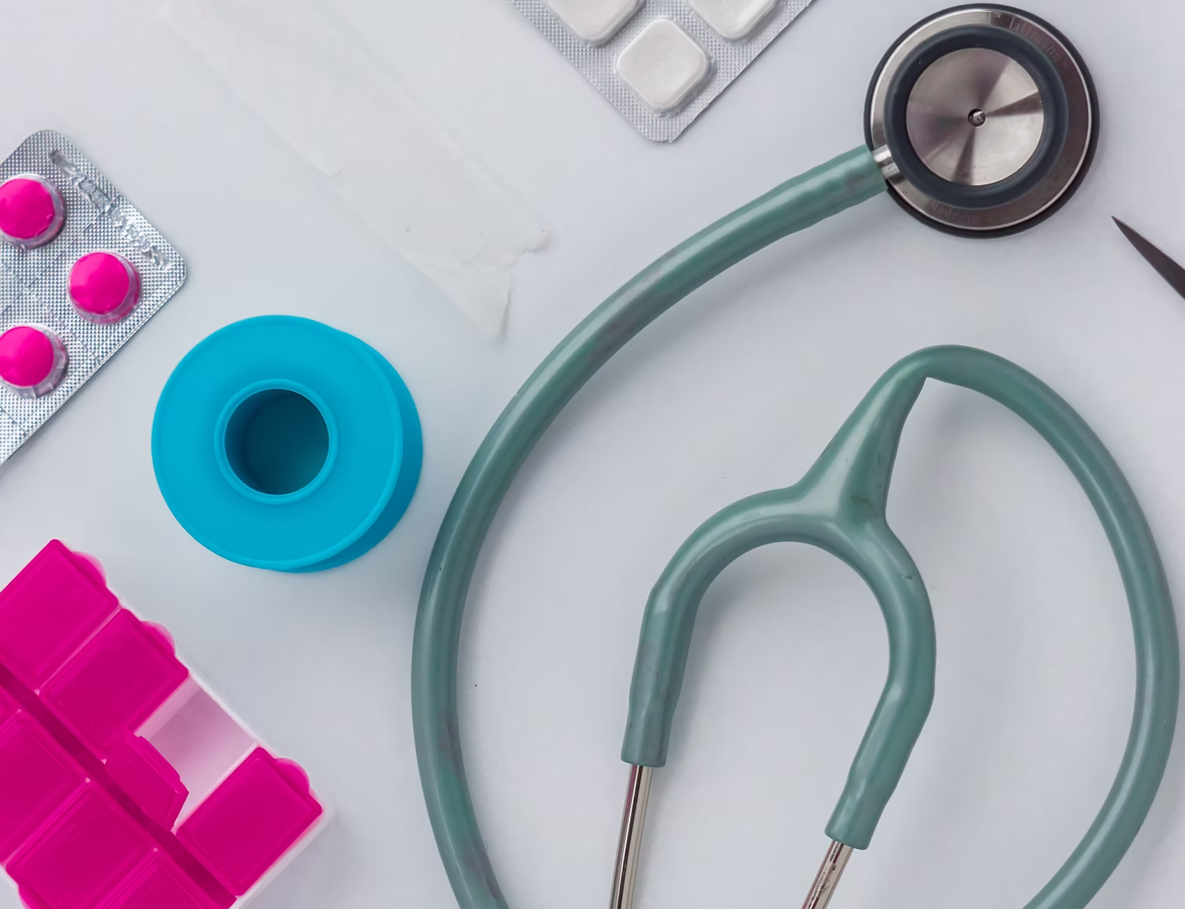 Medical equipment including a stethoscope, blister pack with pink pills, blue silicone container, pink pill organizer, and a pair of scissors on a white surface.