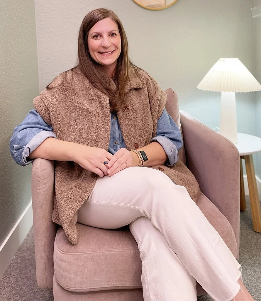 A woman with long brown hair smiling and sitting on a pink armchair, wearing a denim shirt layered with a brown fleece vest, white pants, and a smart watch, in a cozy room with a table lamp.