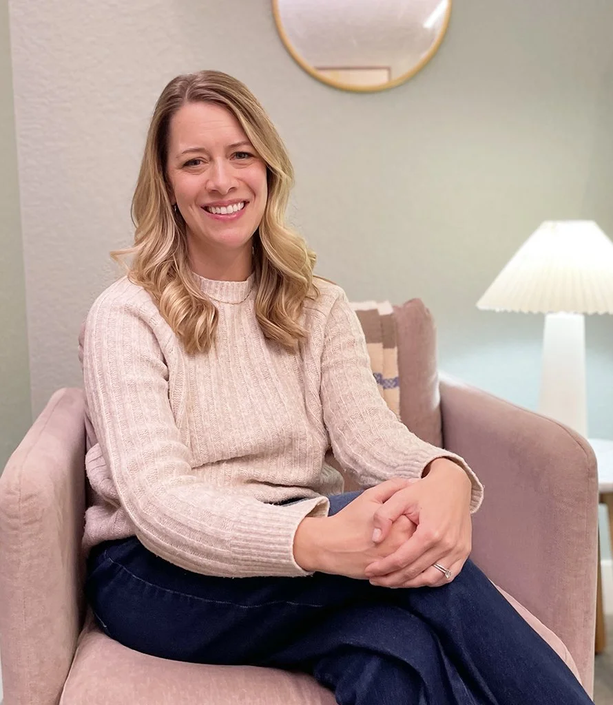 A woman with blonde hair smiling, sitting on a pink armchair in a cozy, softly lit room. She is wearing a light beige sweater and dark blue pants.