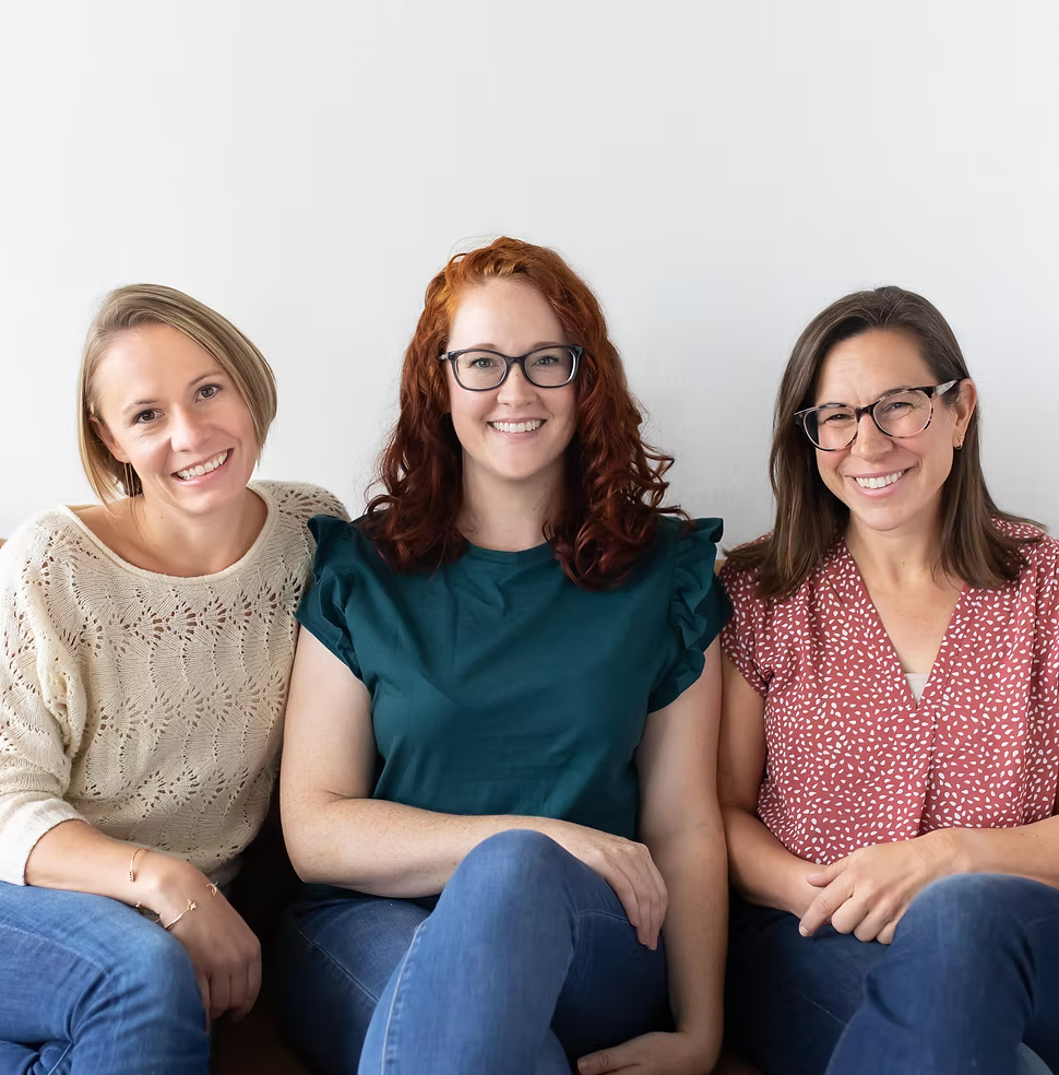 Three women sitting together on a couch, smiling at the camera with a plain white wall in the background.