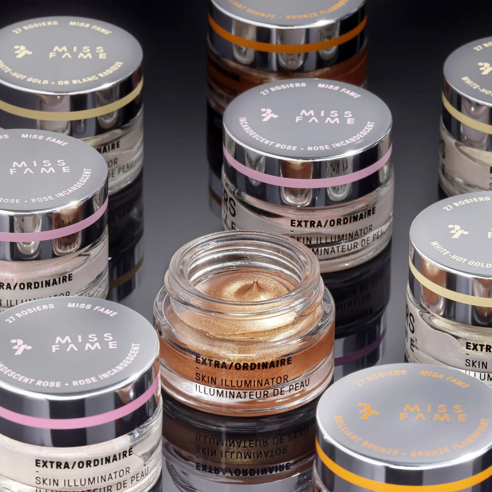 Numerous jars of Miss FAME Skin Illuminator, in various shades, with one open jar showing a golden cream inside. The jars have silver lids with colorful bands and labels.