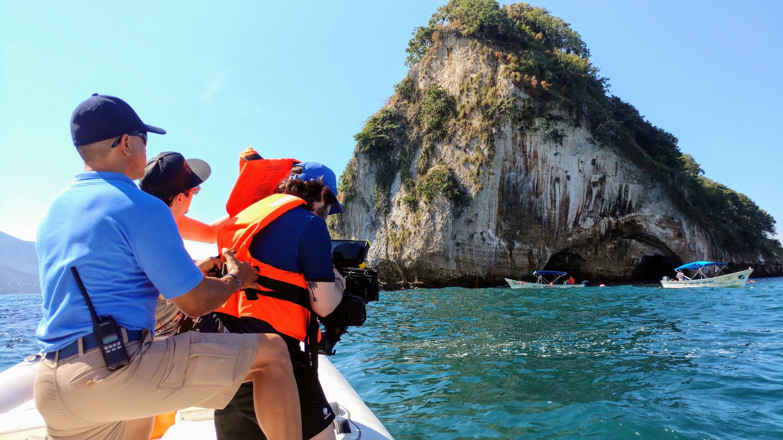 Tourists on a boat near a large sea cave, with a rocky cliff covered in greenery in the background.