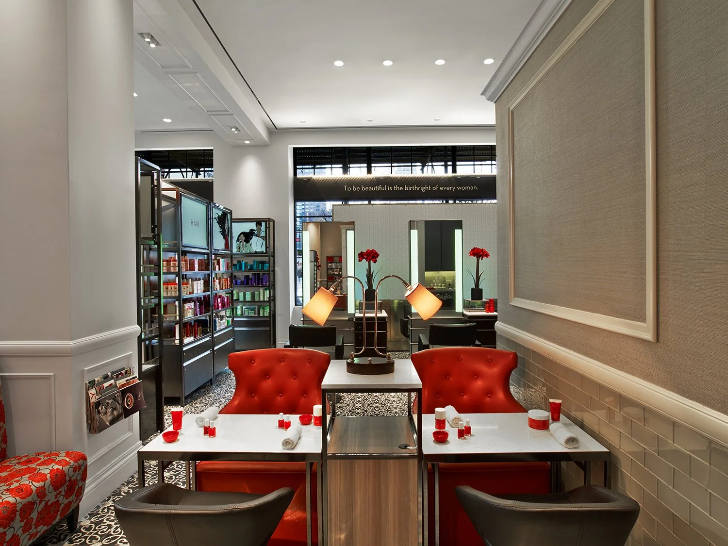 Interior of a restaurant or cafe with red leather chairs, tables set with plates, napkins, and salt and pepper shakers, and shelves with hair products in the background. There are decorative lamps and flowers on the tables.