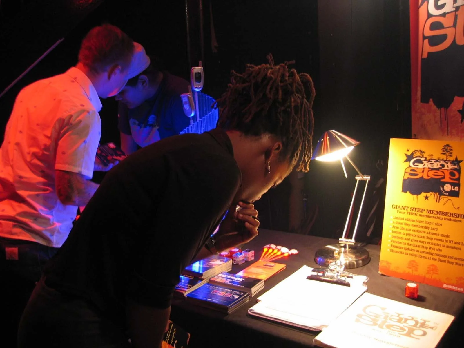 A man with dreadlocks looks at a booth table with flyers, a notebook, and a desk lamp, while two other people stand nearby, examining the materials. There is a bright yellow sign promoting a giant step membership, with additional items on the table.