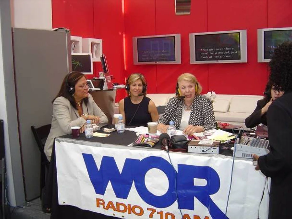Four women wearing headphones sitting at a table with a WOR Radio banner, microphones, and water bottles, engaged in a discussion inside a red-walled studio with screens on the wall.