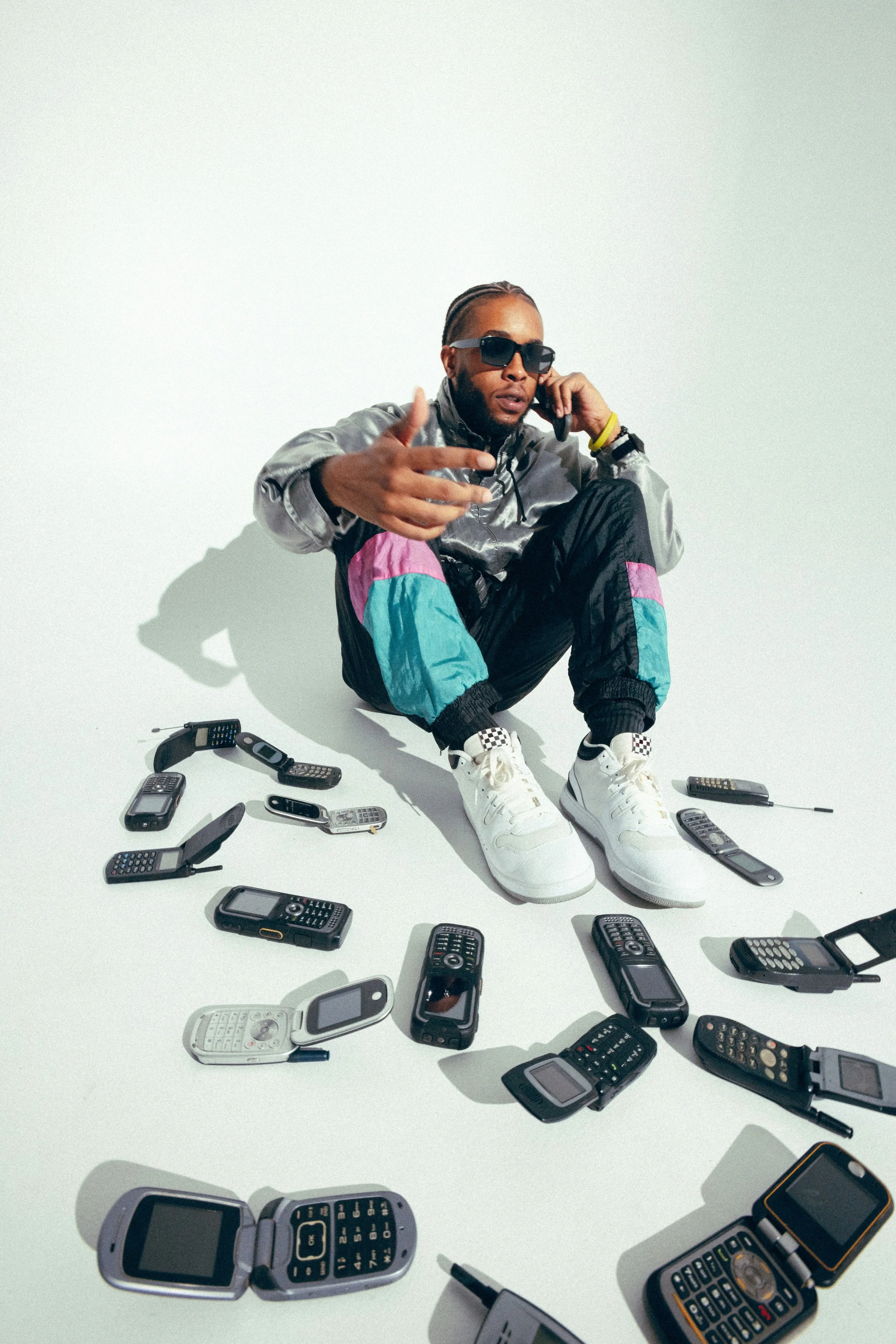 A man wearing sunglasses and colorful retro style clothing sitting on the floor surrounded by numerous old mobile phones.