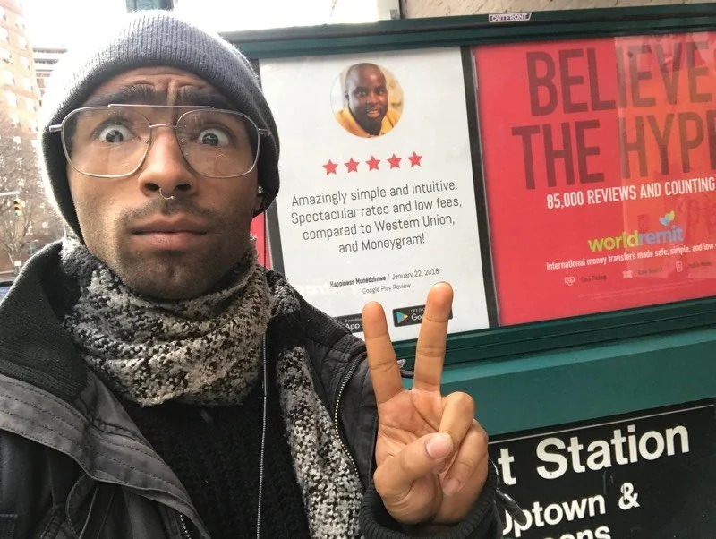 A man making a peace sign taking a selfie near a bus stop with advertisements in Manhattan, New York City.