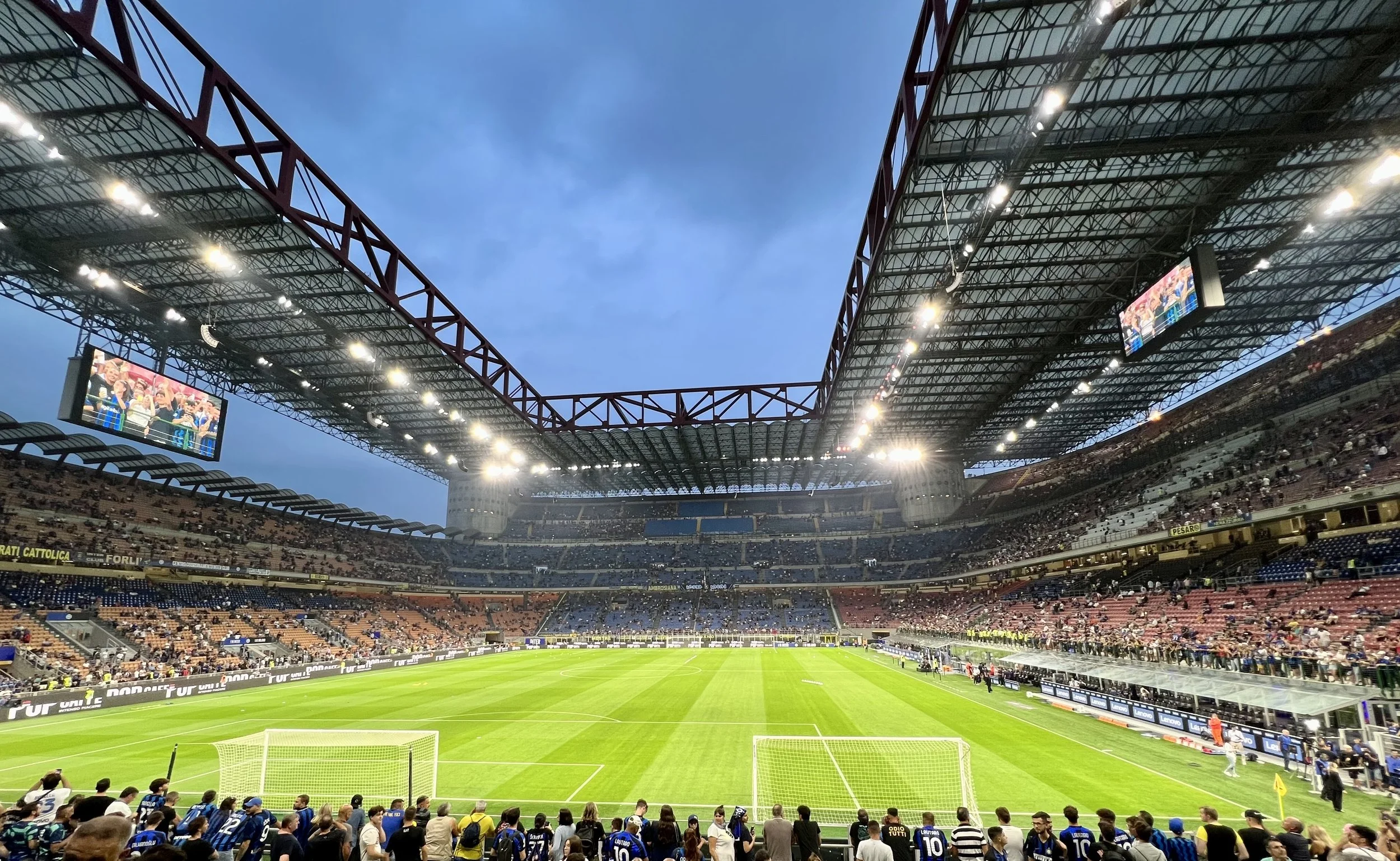 Empty soccer stadium with green field and stands filled with fans, during evening with cloudy sky.