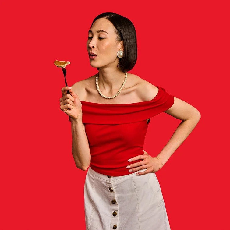 A woman in a red off-the-shoulder top, wearing pearl earrings and necklace, holding a fork with a lemon wedge, against a red background.