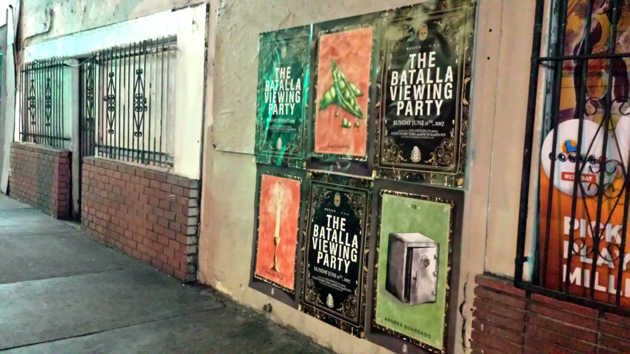 A sidewalk with a wall displaying multiple posters about 'The Batalla Viewing Party' event on June 11, 2017. The wall has a brick and plaster surface, with a window barred by black decorative metal grills and another poster on a metal grid.