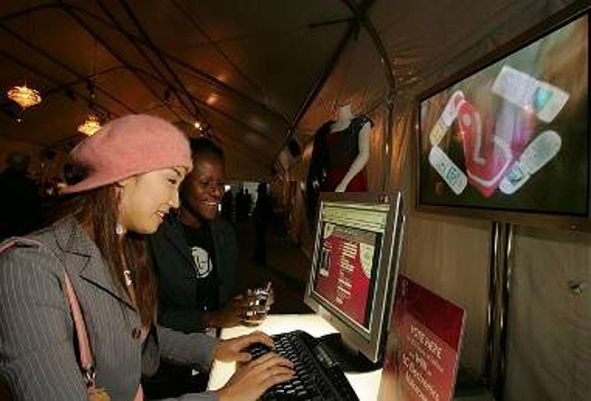Two women at a shopping mall browsing information on a computer screen, with promotional posters and mannequins displaying clothing in the background.