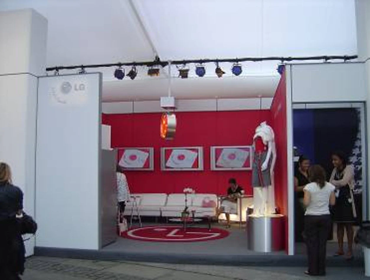 An indoor event booth with a red and white theme, featuring a sofa, three framed artwork pieces, a Santa hat, and several people standing and sitting inside and outside the booth.