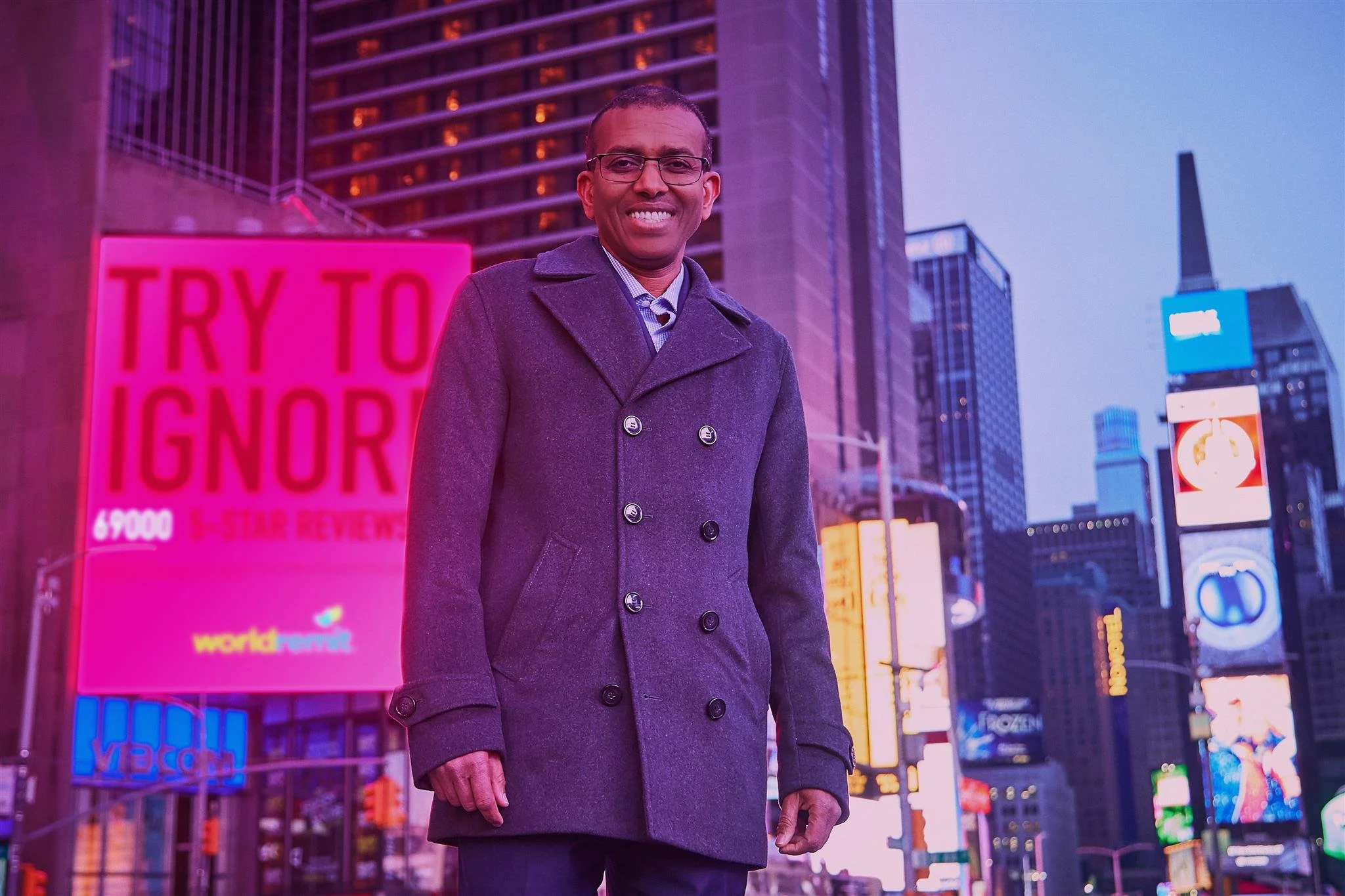 A smiling man in a gray coat standing in Times Square at dusk with illuminated billboards and skyscrapers in the background.