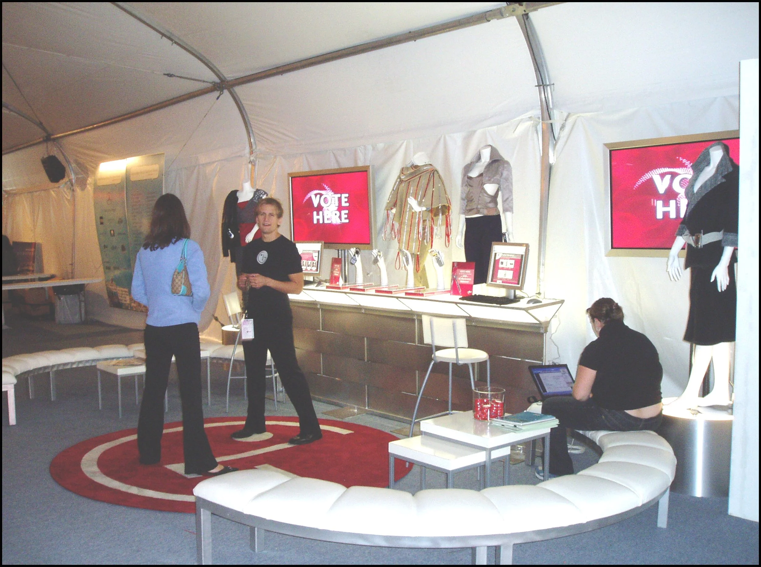 Fashion booth with mannequins dressed in clothing, digital screens displaying 'Vote Here,' two women talking, one woman seated on a curved white bench working on a laptop.