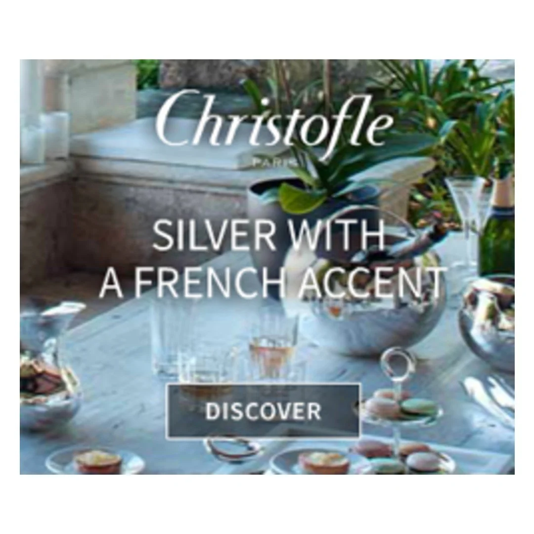 A table with silver bowls and glasses, potted plants, and a window in the background. Overlaid text reads "Christofle Paris Silver with a French Accent" and a button labeled "Discover."