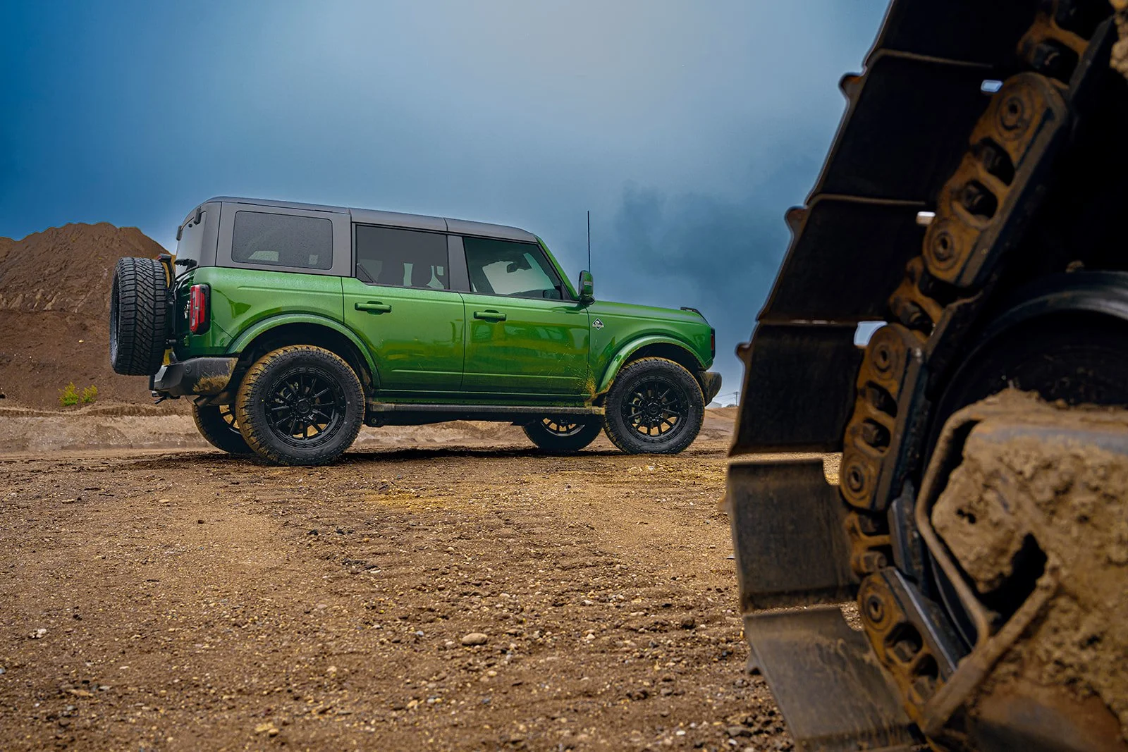 A green off-road vehicle parked on a dirt terrain with a construction or excavation vehicle track in the foreground and a cloudy sky in the background.