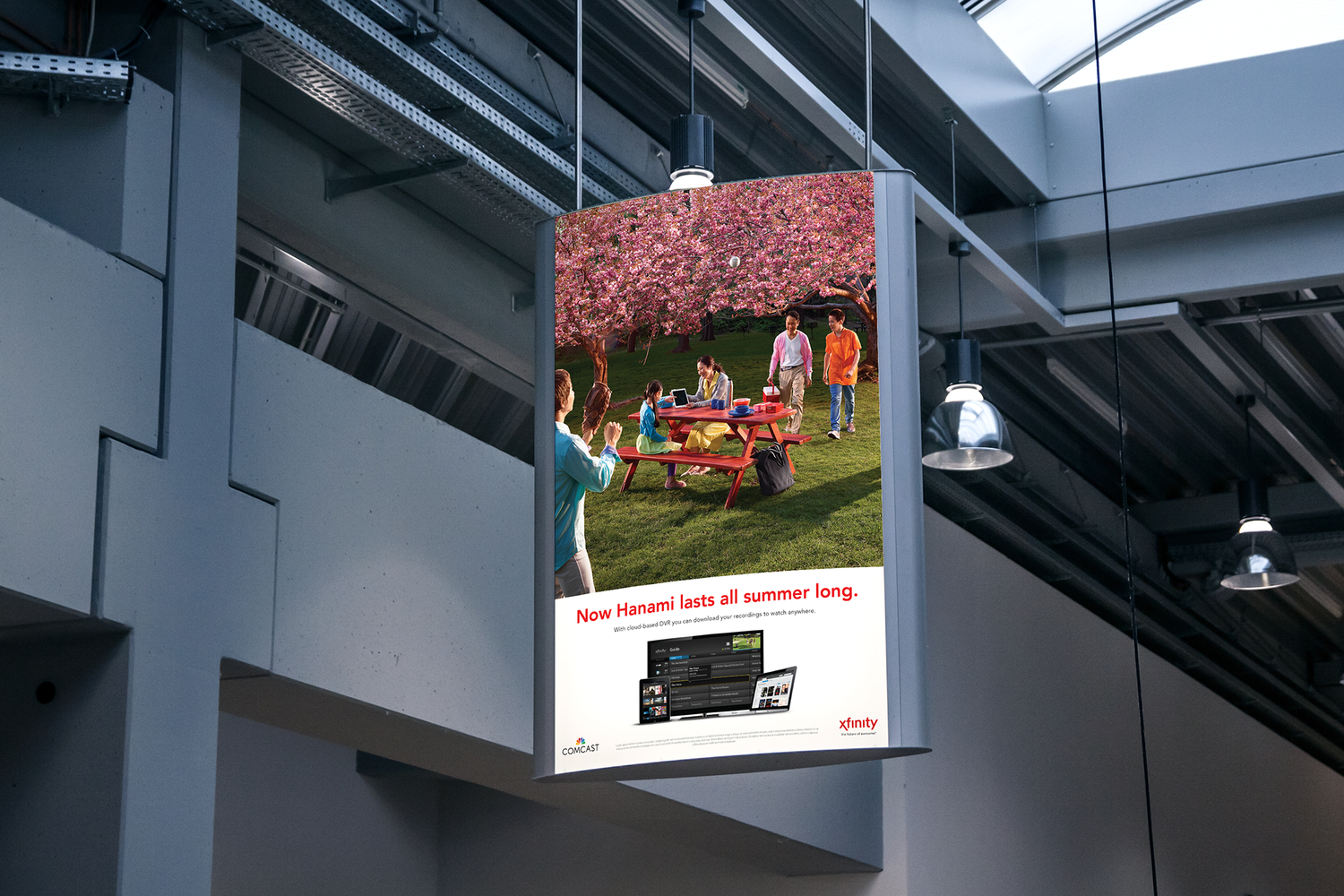 An advertisement hanging from the ceiling of a modern building showing a group of people enjoying a picnic under cherry blossom trees outside. The lower part of the ad promotes Xfinity's cloud-based DVR service with images of devices and the text, 'Now Hanami lasts all summer long.'