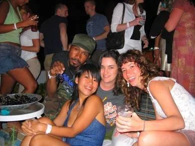 Four friends smiling and posing for a photo at a crowded indoor event, with drinks in their hands.