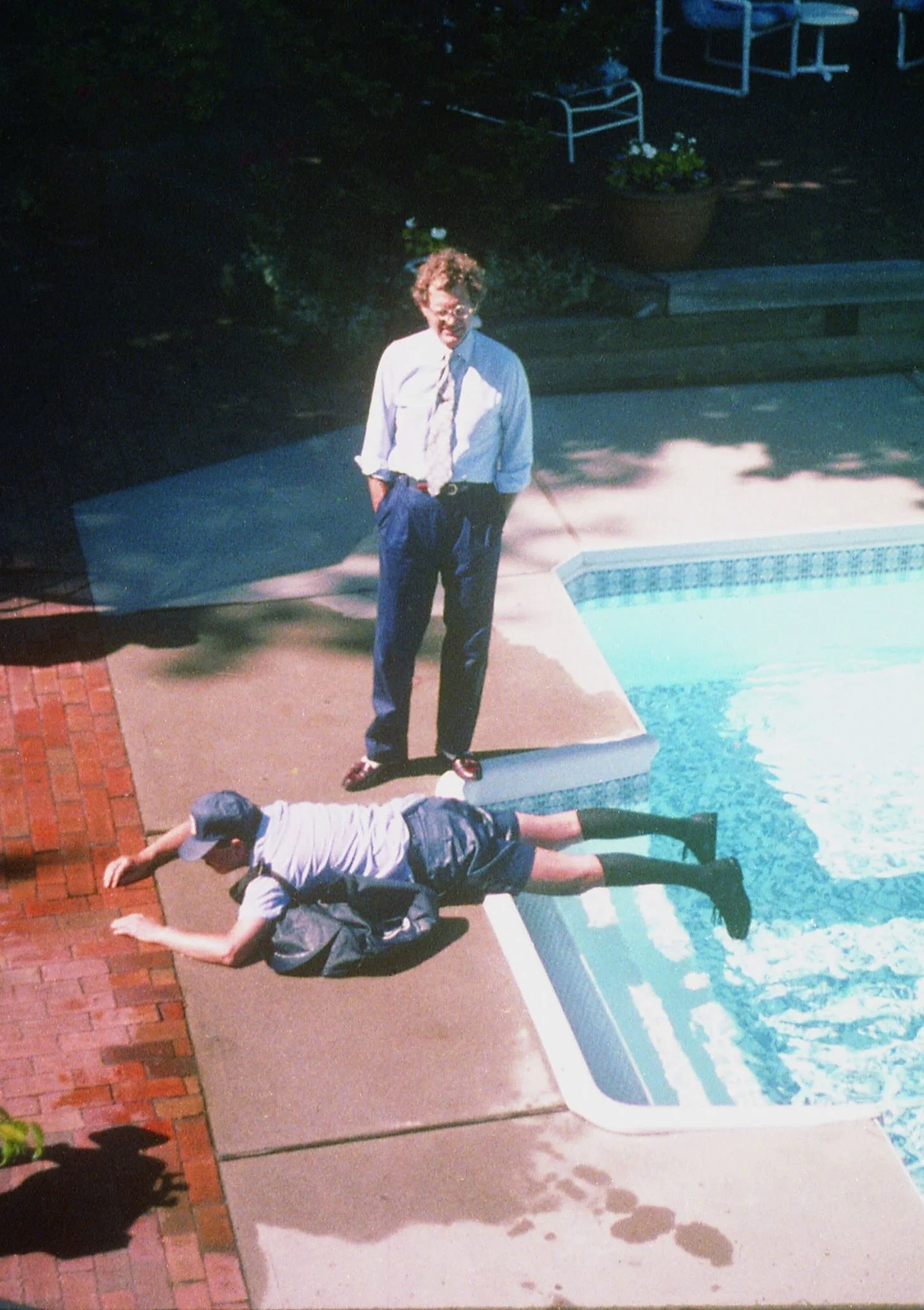 A photo of two children by a swimming pool; one is lying on the ground and the other is standing nearby with hands in pockets, dressed in a white shirt, tie, and blue pants.