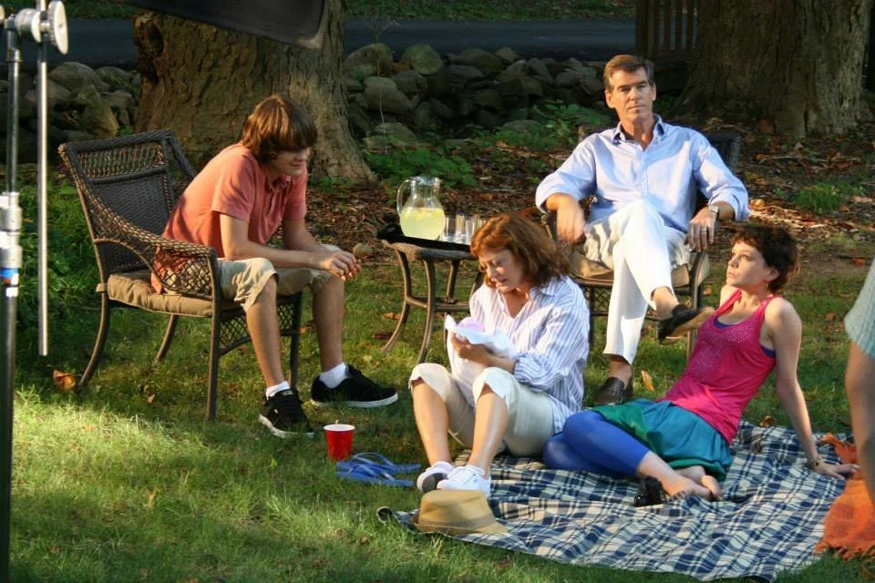 A group of five people, three women and two men, are gathered outdoors during daytime, sitting on chairs and a blanket on the grass, enjoying a relaxed social gathering or picnic near trees and rocks.
