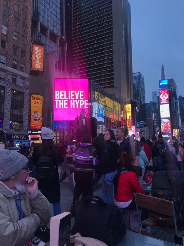 People gathered in Times Square at night, illuminated by bright neon billboards, with a large pink LED sign reading "BELIEVE THE HYPE" and a metal sculpture of a car in the foreground.