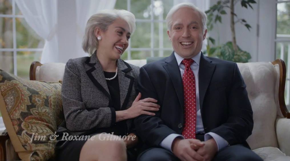An older man and woman sitting on a sofa, smiling and laughing. The woman has gray hair and the man has white hair, both dressed in formal attire. They're in a bright room with large windows and greenery outside.