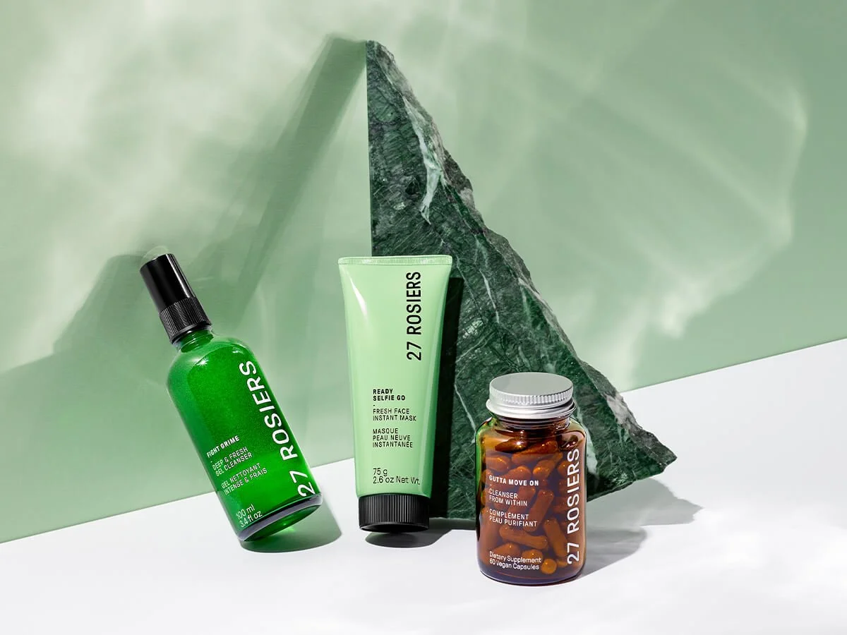 Green spray bottle, green face mask tube, and brown supplement bottle with capsules, all from 27 Rosiers, arranged on a white surface with a green stone block and green background.