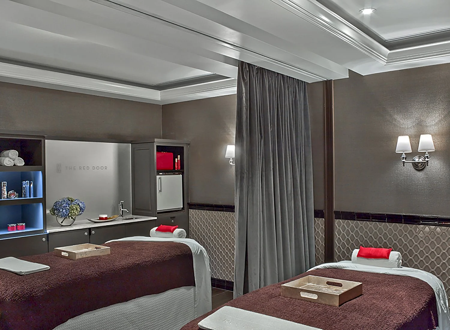 A hotel massage room with two massage tables covered in burgundy blankets, each with a pillow and rolled towel. There is a small tray on each table. The room has beige walls, elegant light fixtures, and blue bookshelves with towels and decorative items. A curtain separates the space.