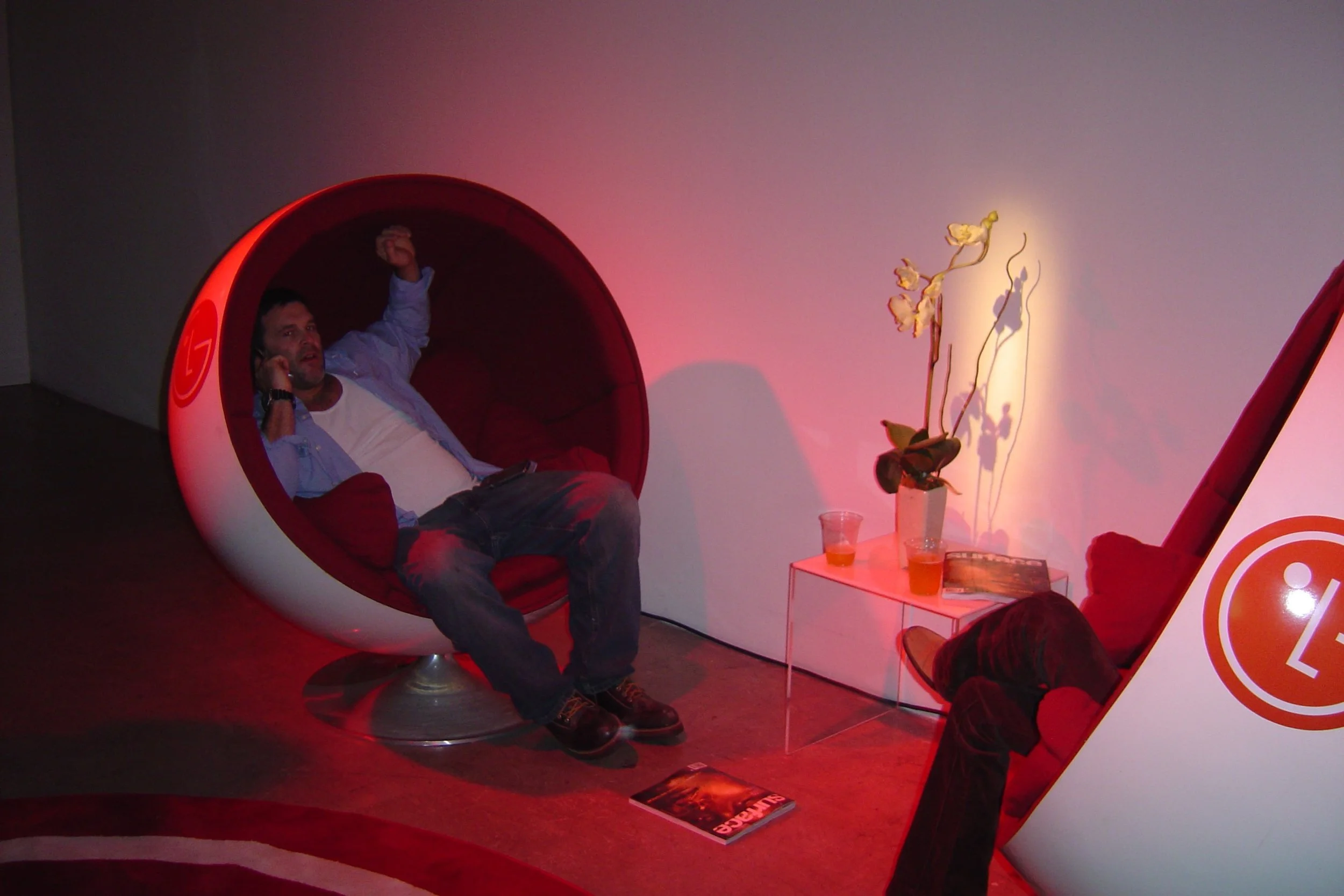 Two men are sitting in modern red and white spherical chairs in a dimly lit room, engaging in phone conversations. A small, transparent table holds drinks and a magazine, with a potted orchid casting shadows on the wall behind them.