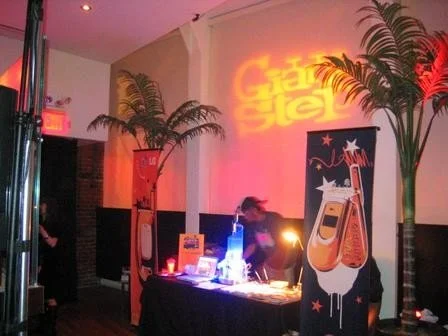 Indoor event space with tropical plants, a table with promotional materials, and a person working behind it. Neon sign on the wall reads "Greatest Glo" in red and orange.