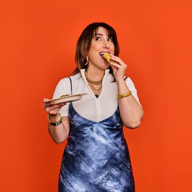 A woman with shoulder-length brown hair, wearing a white top and blue dress, is eating a snack from a plate she is holding in her left hand, smiling and looking to the side against an orange background.