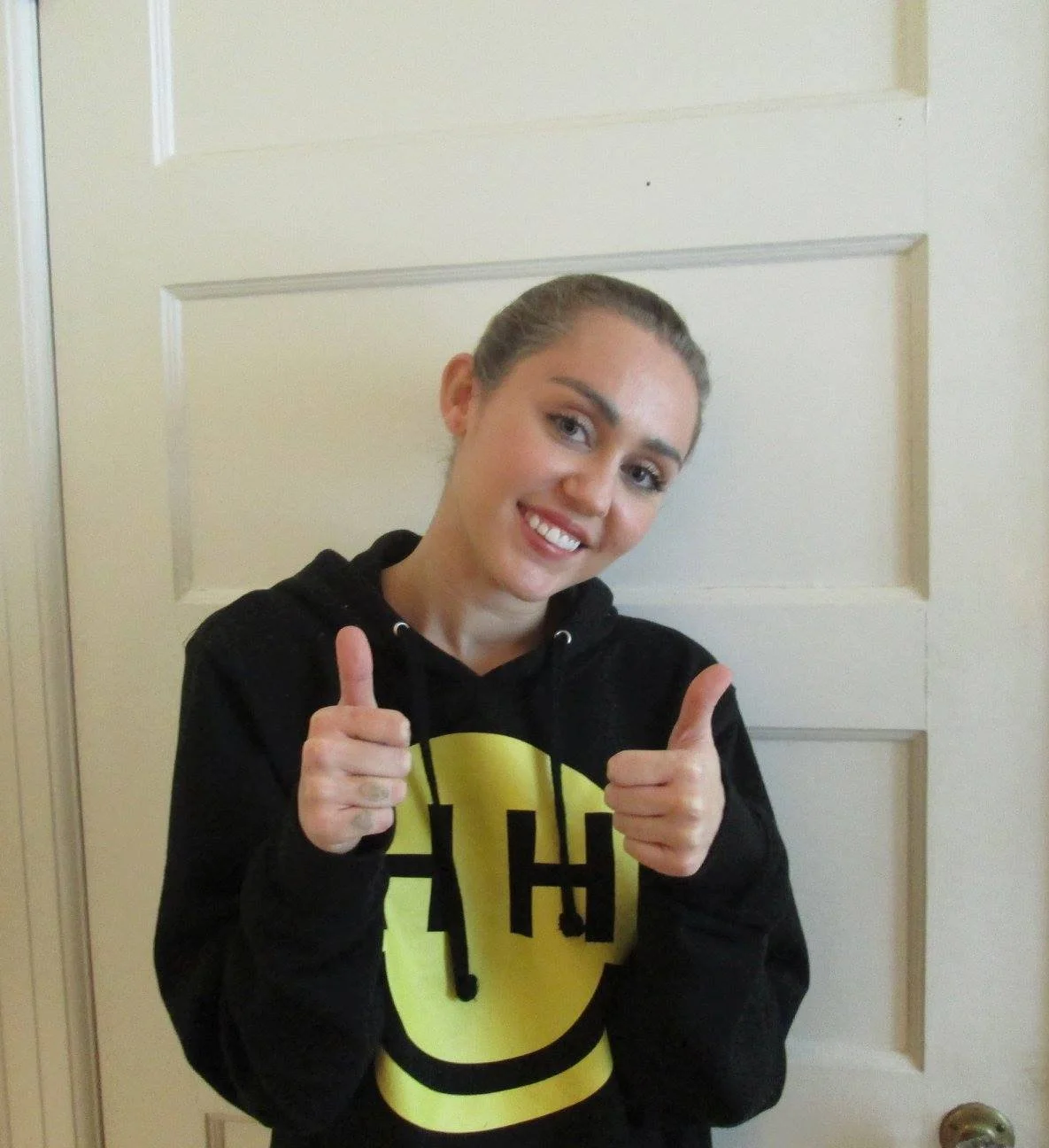 A young woman with braided hair smiling and giving a thumbs-up gesture, wearing a black hoodie with a yellow smiley face logo.
