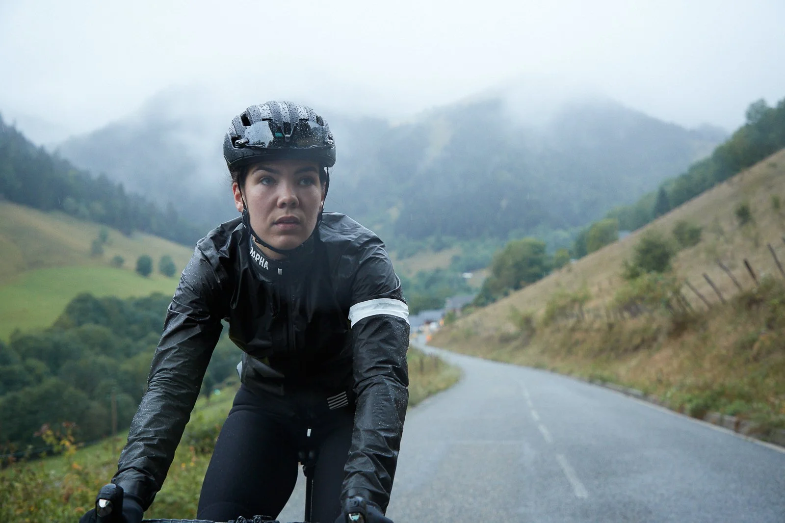 Woman riding a bicycle on a rainy mountain road, wearing a black helmet and rain jacket, with hills and trees in the background.