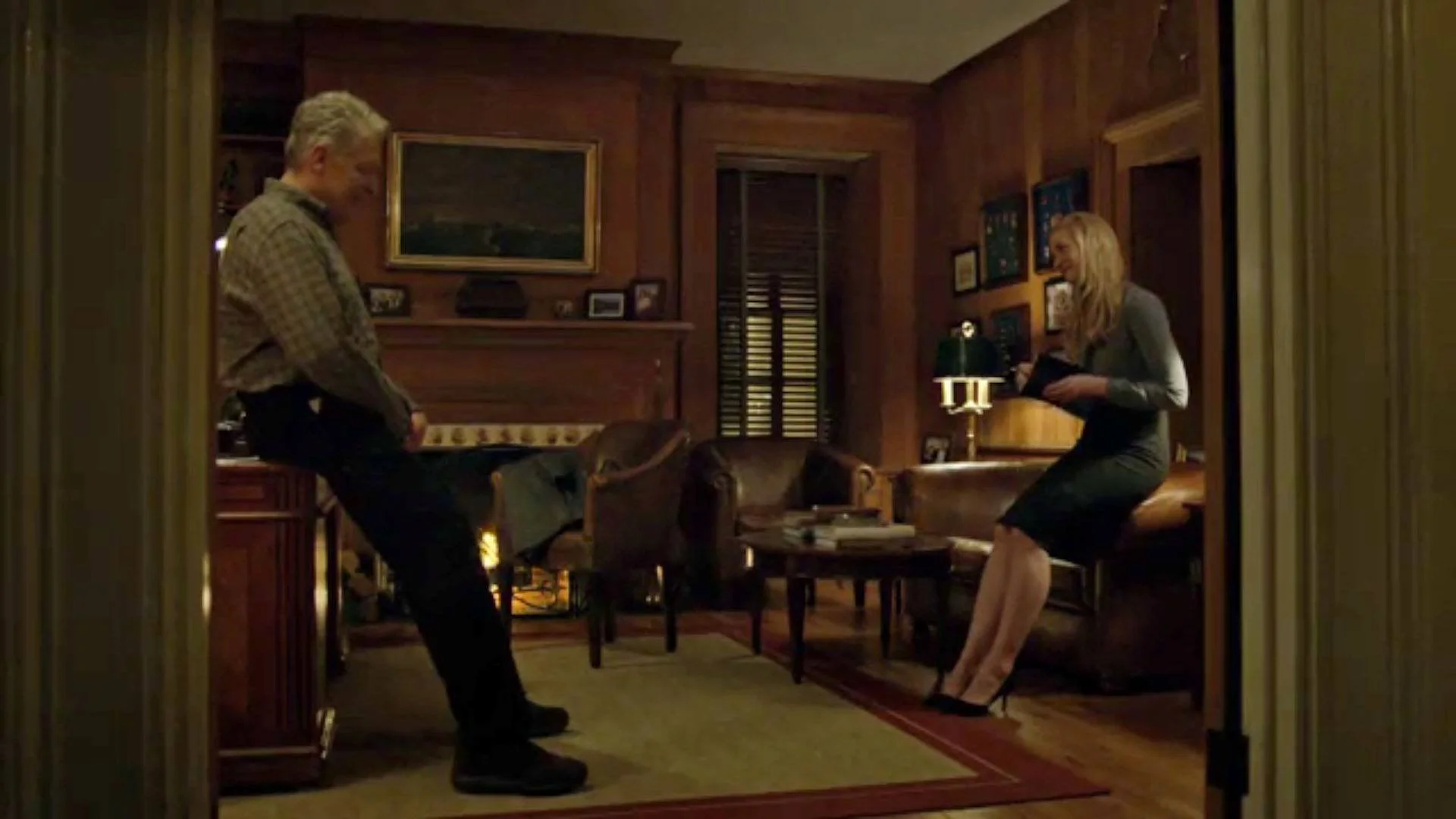 A man and a woman in a warmly lit, wood-paneled living room, standing opposite each other and engaging in a conversation. The man is leaning against a cabinet, while the woman is standing on heels holding a book or folder.