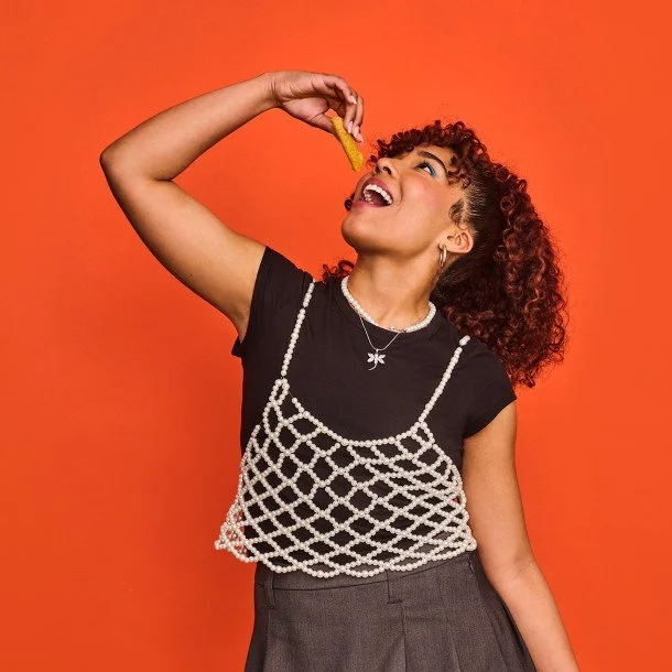 A young woman with curly hair wearing a black t-shirt and gray skirt, smiling as she holds a fry above her mouth against an orange background.