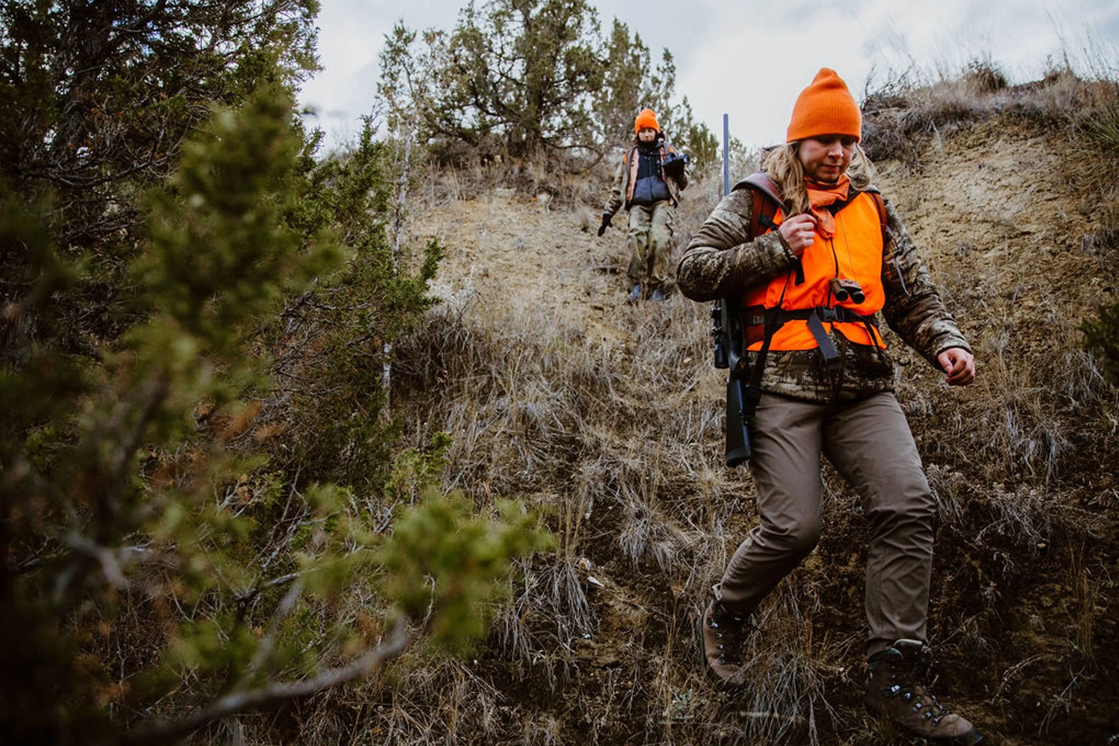 Two people hiking downhill on a rugged trail in a dry, wooded area, both wearing orange hats and backpacks.
