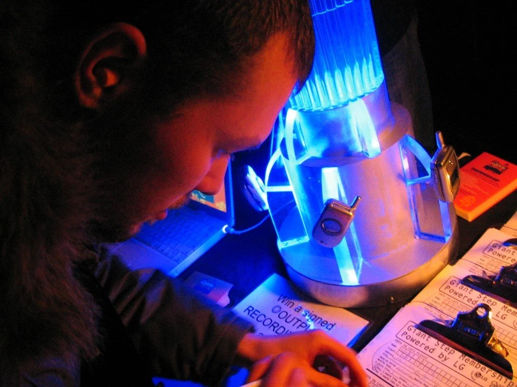 Person looking at a device with a large glass apparatus illuminated in blue light, surrounded by paperwork and electronic equipment.