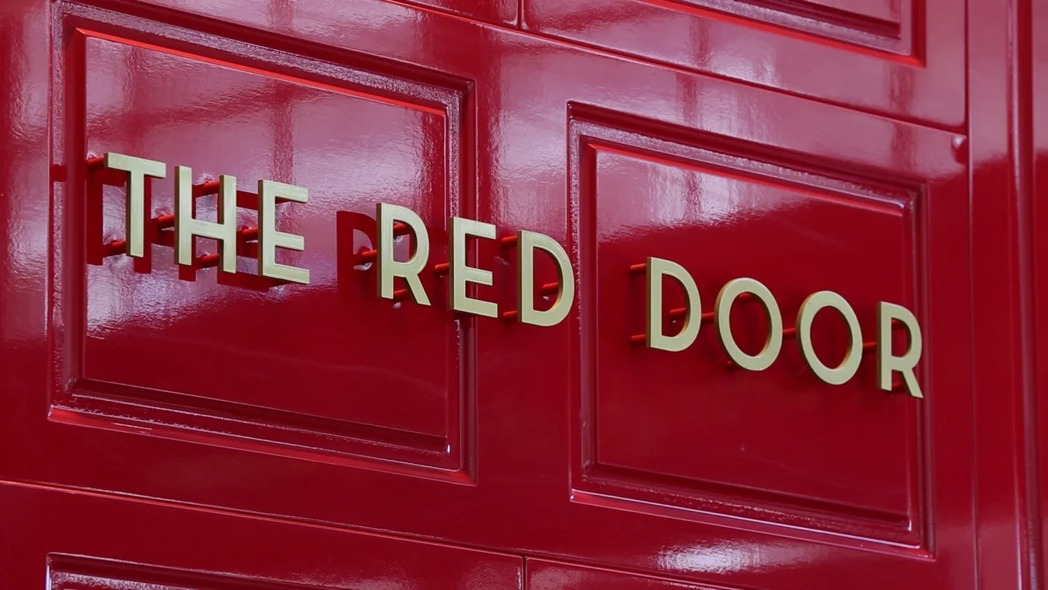 Close-up of a shiny red door with raised panel design and a sign that says "The Red Door" in white 3D lettering.