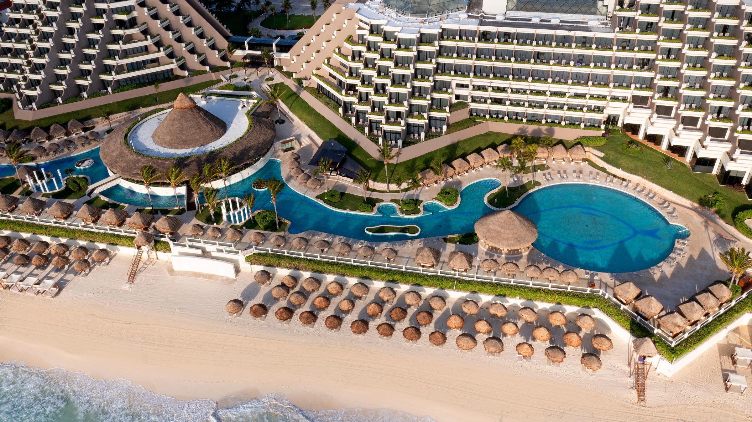 Aerial view of a beachfront resort with infinity pools, palm trees, and sun umbrellas on the sandy beach.