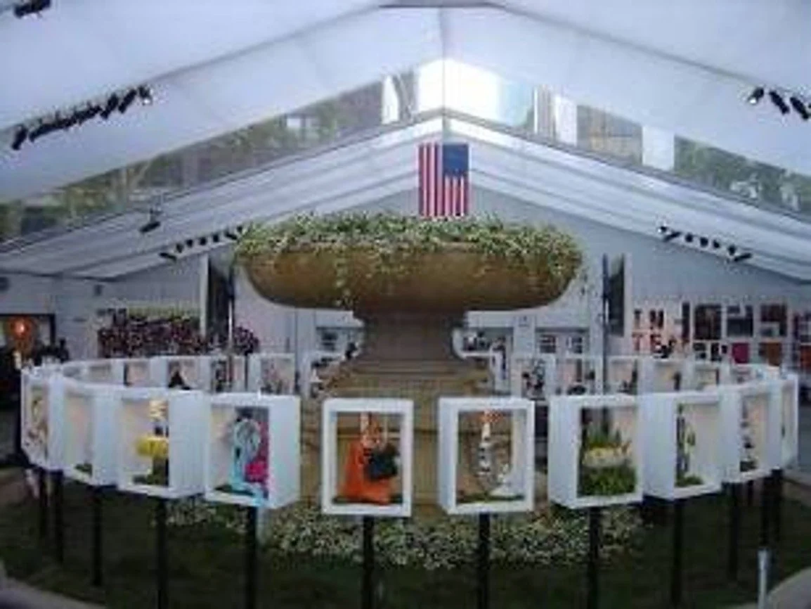 An indoor gallery exhibit featuring artwork displayed in white frames arranged in a circle around a large stone planter with green plants. An American flag hangs above the display inside a glass-roofed structure.