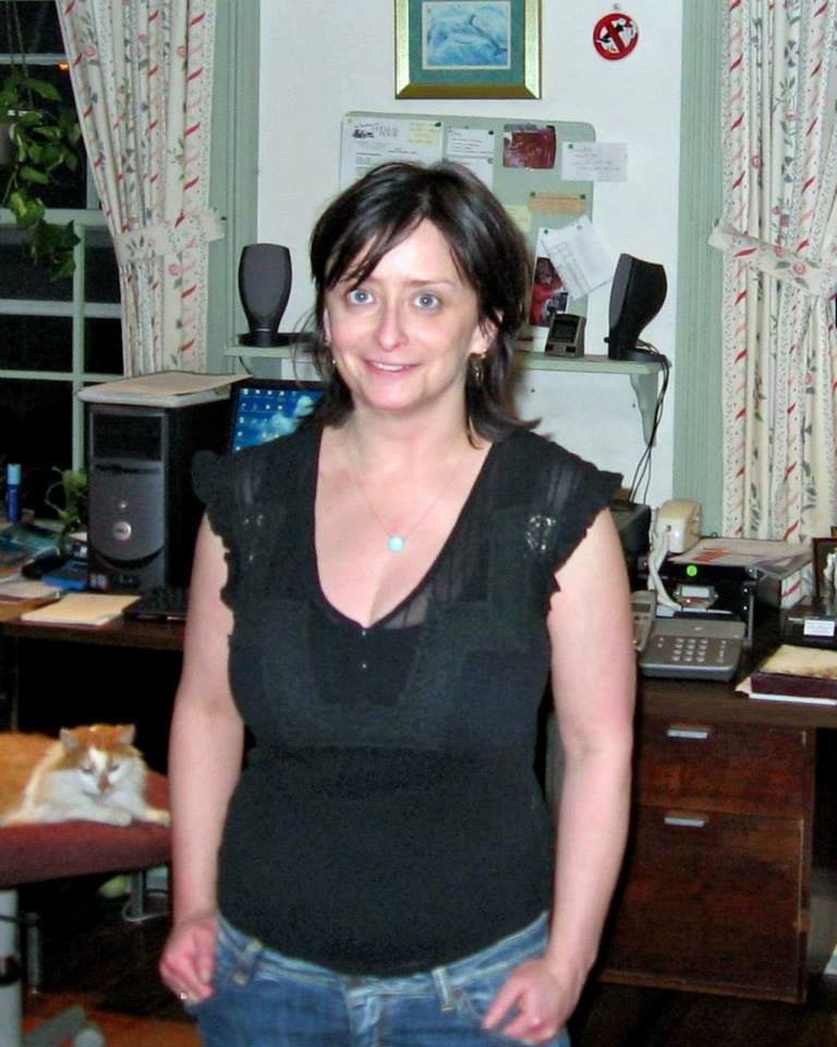 A woman standing in an office with computer equipment and a calico cat sitting on a chair behind her.