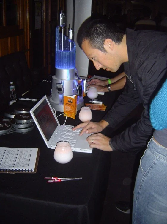 A man leaning over a white laptop at a table with various electronic devices, drinks, and a blue fountain on the table.