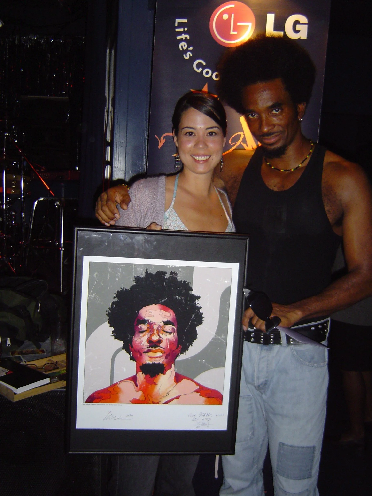 Two people standing together, holding a framed digital art portrait of a man with afro hairstyle and a goatee, in front of an LG sign with the slogan "Life's Good."