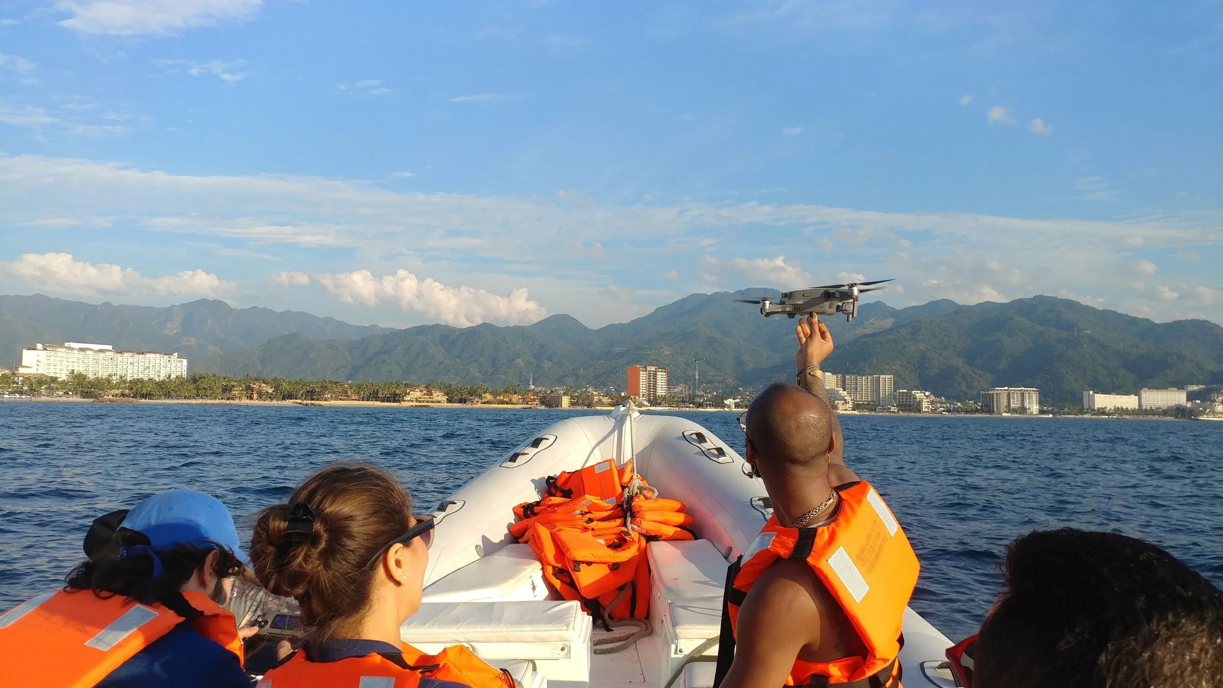 People in life jackets on a boat, with a man holding a drone, on a body of water with mountains and buildings in the background.