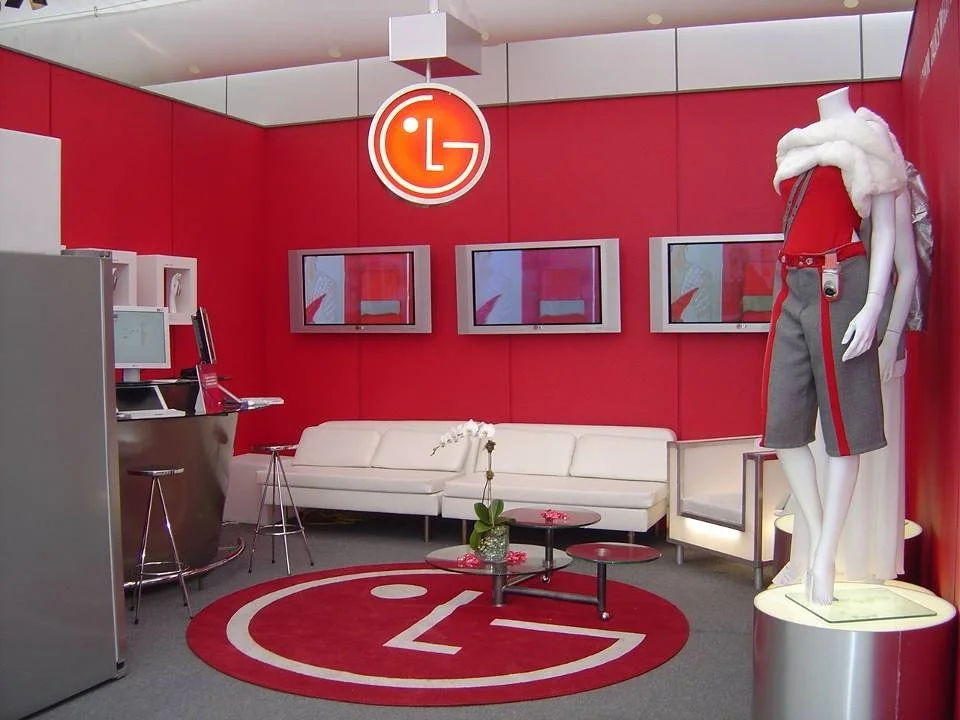 A modern LG showroom with a red wall, a white sofa, four wall-mounted screens, a mannequin dressed in casual clothes, and an LG logo hanging from the ceiling.