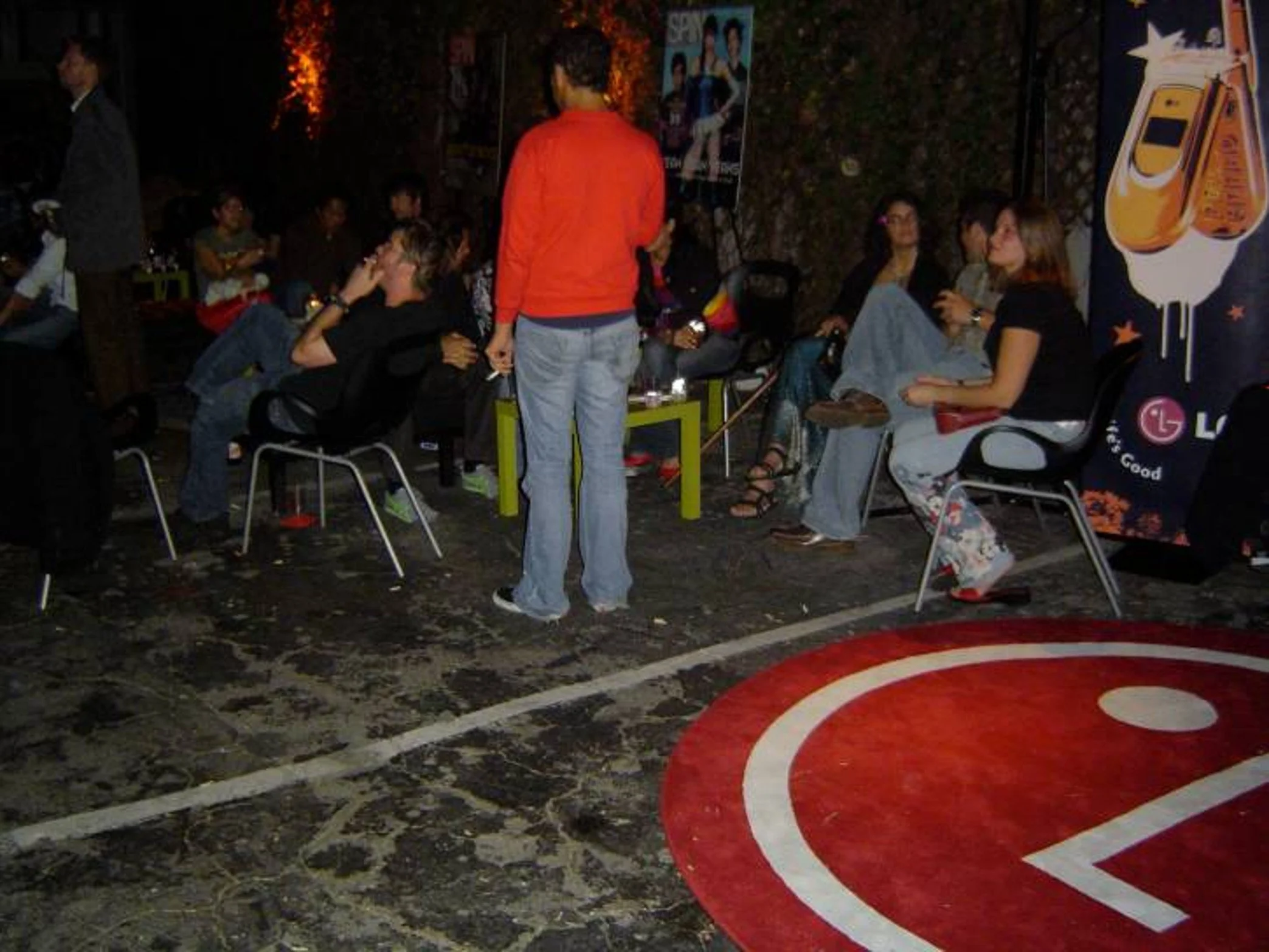 Group of people sitting and standing at an outdoor gathering at night. Some are seated on chairs, engaged in conversation, while one person in a black shirt sits on a chair with floral pants. A person in an orange jacket stands in the center, and oth