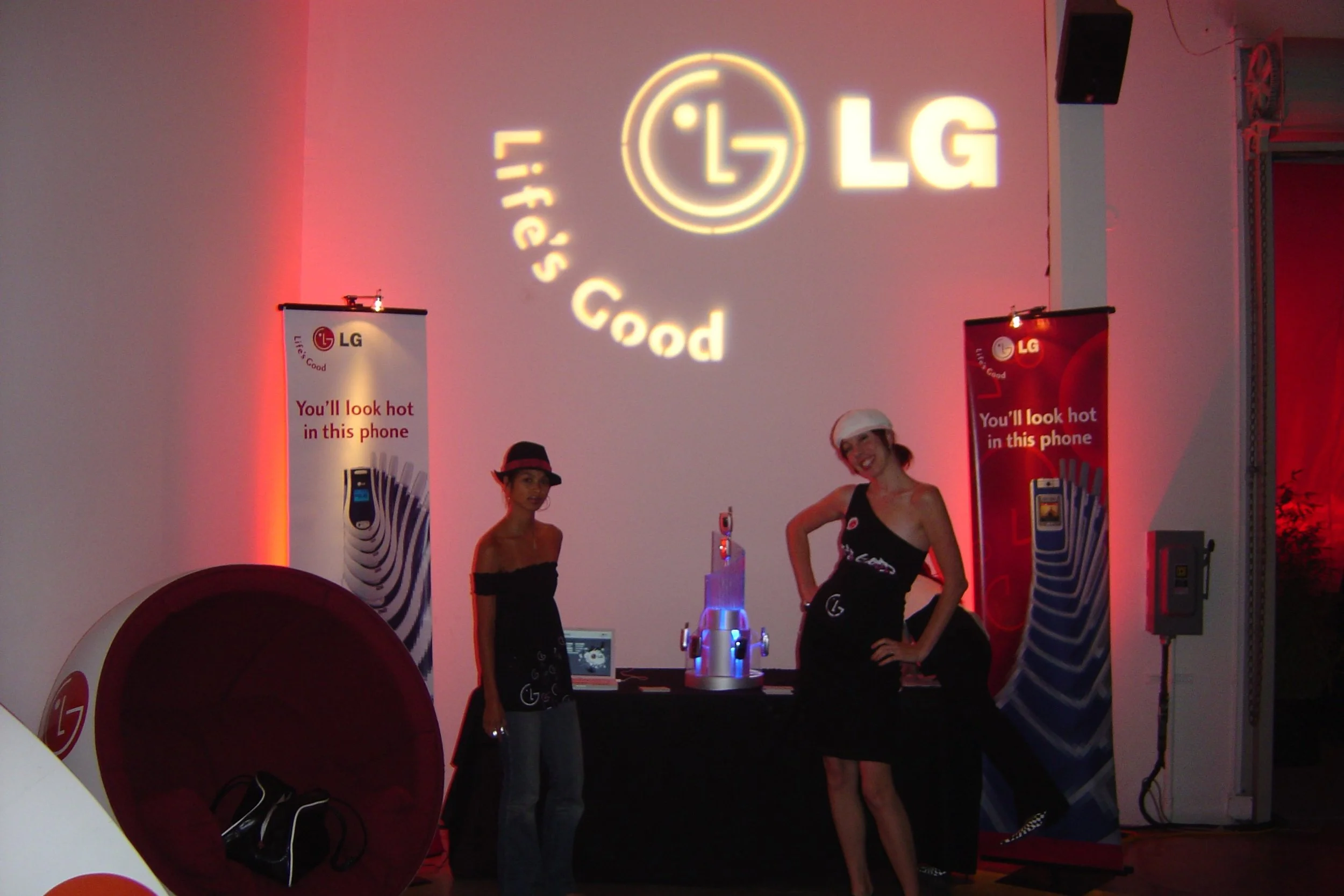 Two women at a promotional event for LG, standing in front of a wall with the LG logo and slogan "Life's Good" projected on it. The woman on the left is wearing a hat, and the woman on the right is wearing a white headband. There are red banners with