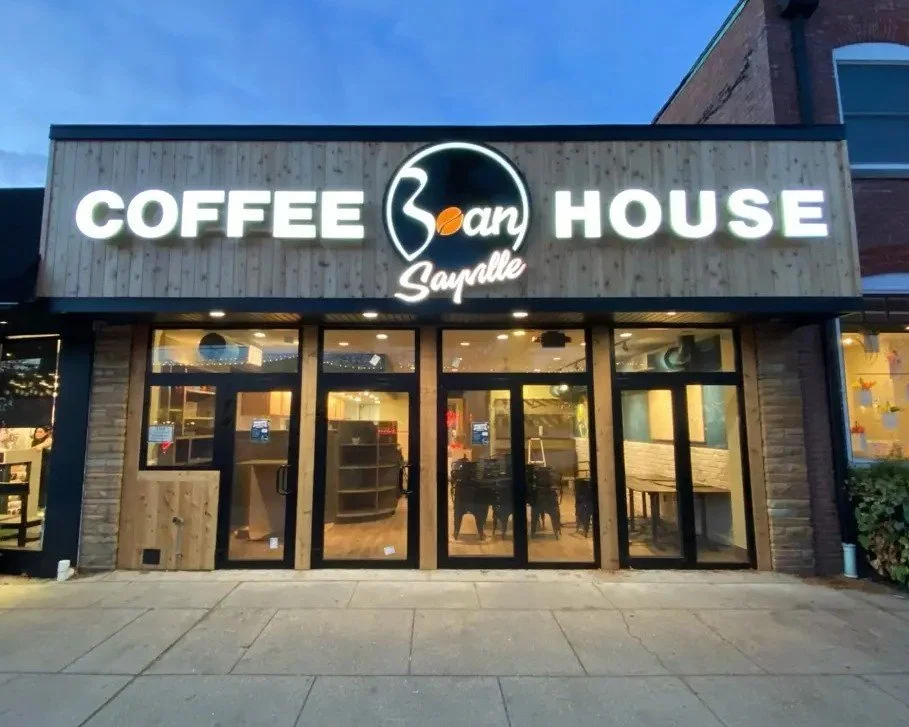 Front view of a coffee shop with illuminated sign reading 'Coffee 3 an House' and 'Sayville' underneath, with glass doors and windows showing the interior.
