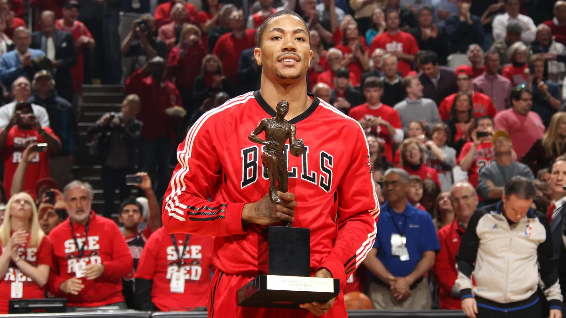 A basketball player wearing a red Bulls jersey holding a trophy in front of a crowd celebrating.