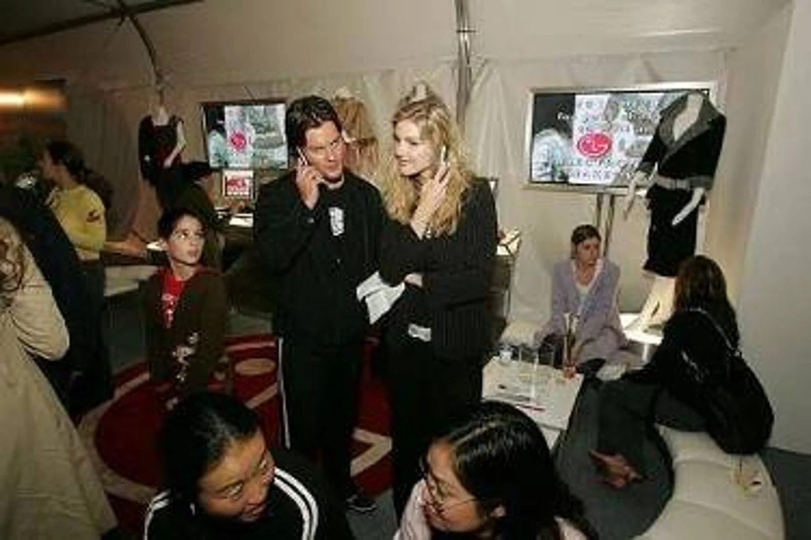 A group of people in a room, some sitting and some standing. Two women are talking on the phone. There are TV screens in the background displaying logos and mannequins wearing clothing.